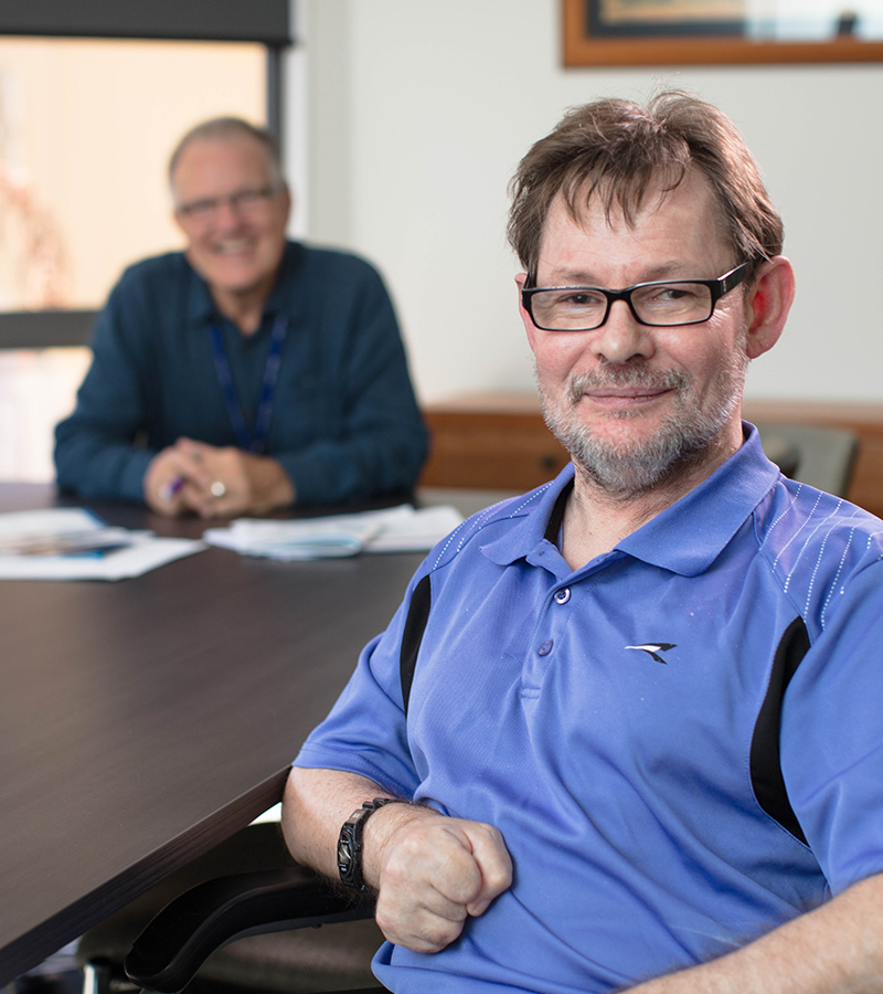 man wearing glasses and a purple shirt smiles while sitting at a desk indoors