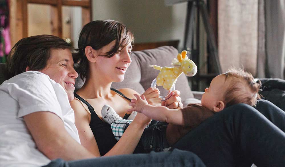 young man and woman sitting on a couch with the woman holding a teddy bear while playing with an infant child resting on her legs