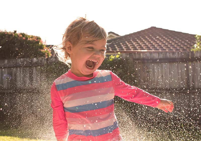 Laughing toddler splashes in a backyard blow-up pool while a woman sprays her with water from the hose.