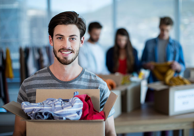 young man holding a box of donated clothes smiling in the foreground with workers packing more boxes of clothes in the background