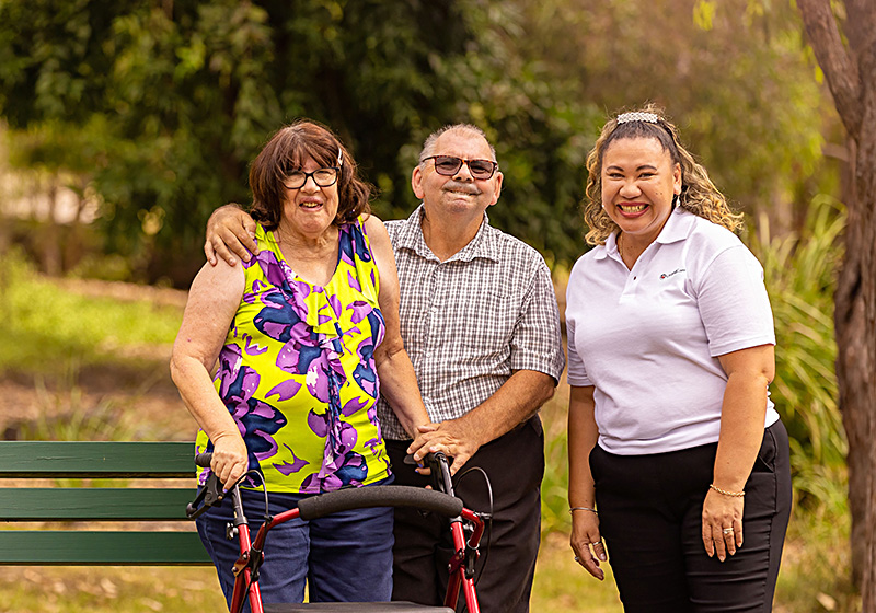 A woman who uses a wheelie walker stands with her partner and a UnitingCare disability support worker beside a bench in a country park.