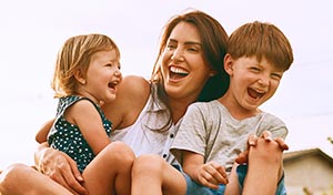 A woman with a young girl and boy in her lap sitting in parklands, laughing and smiling