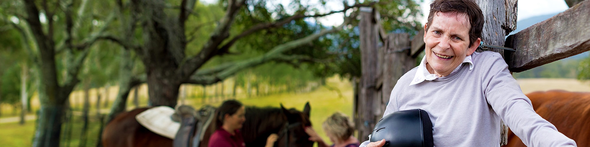 disabled man leaning against fence outdoors with staff attending a horse in the background