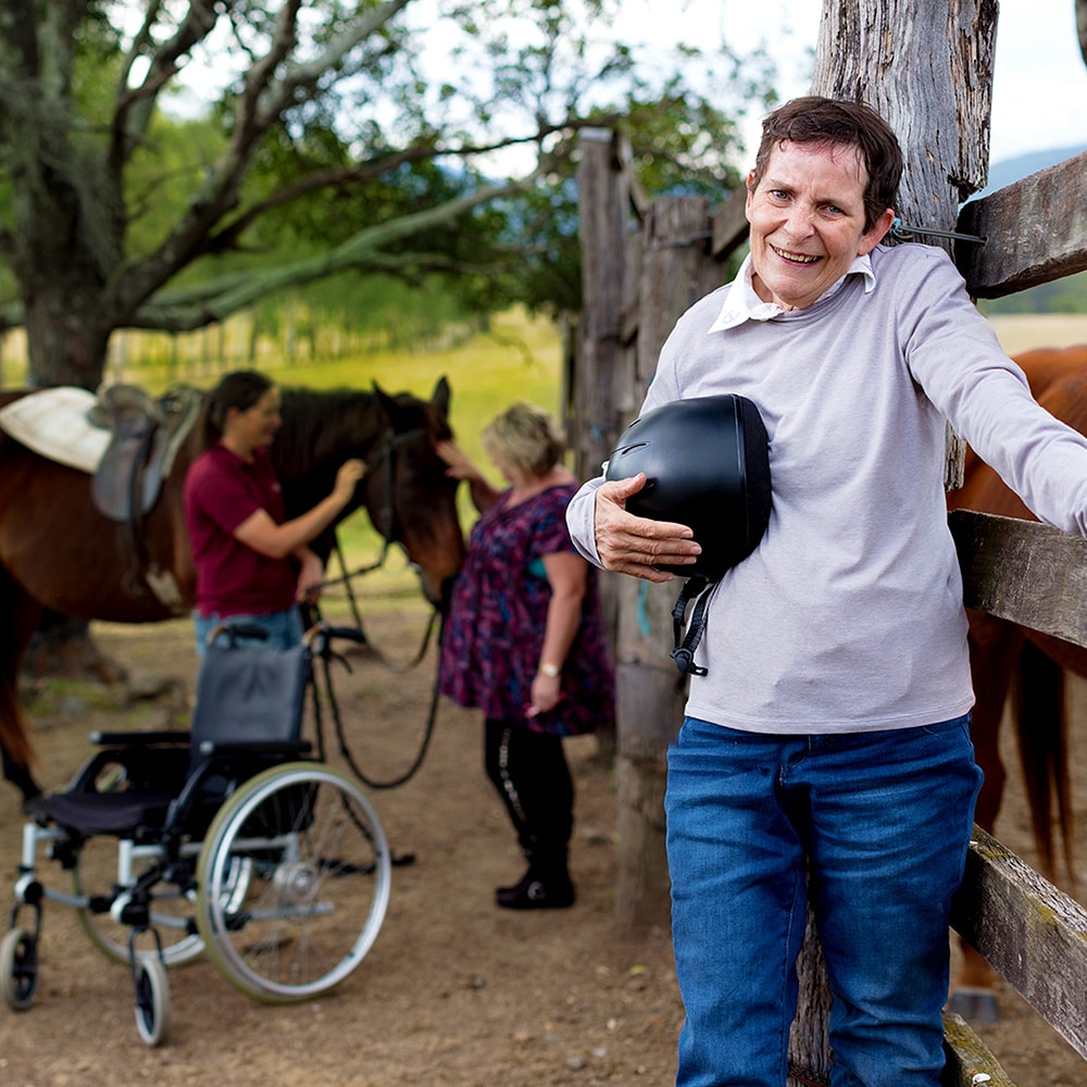disabled man leaning against fence outdoors with staff attending a horse in the background