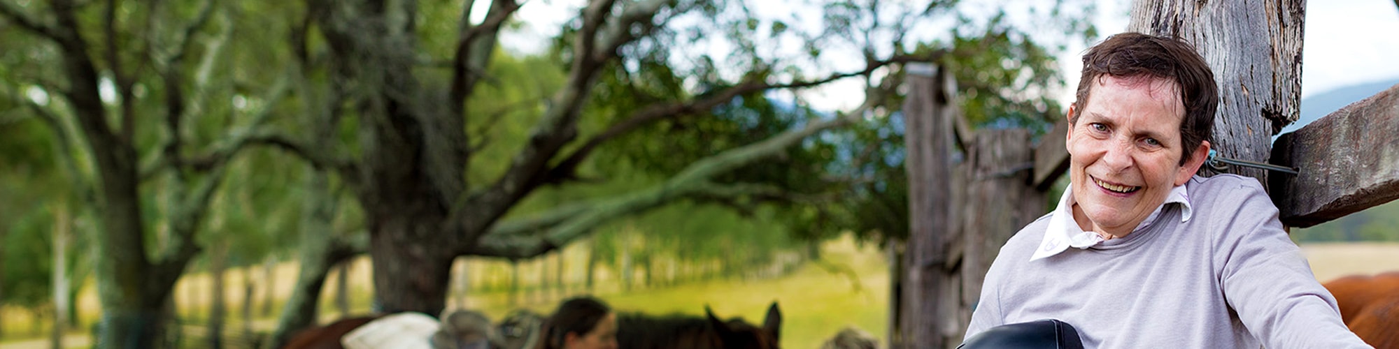 disabled man leaning against fence outdoors with staff attending a horse in the background