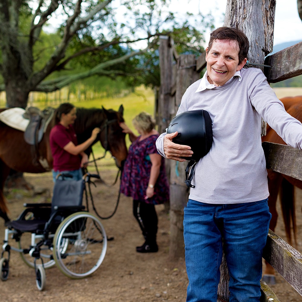 disabled man leaning against fence outdoors with staff attending a horse in the background