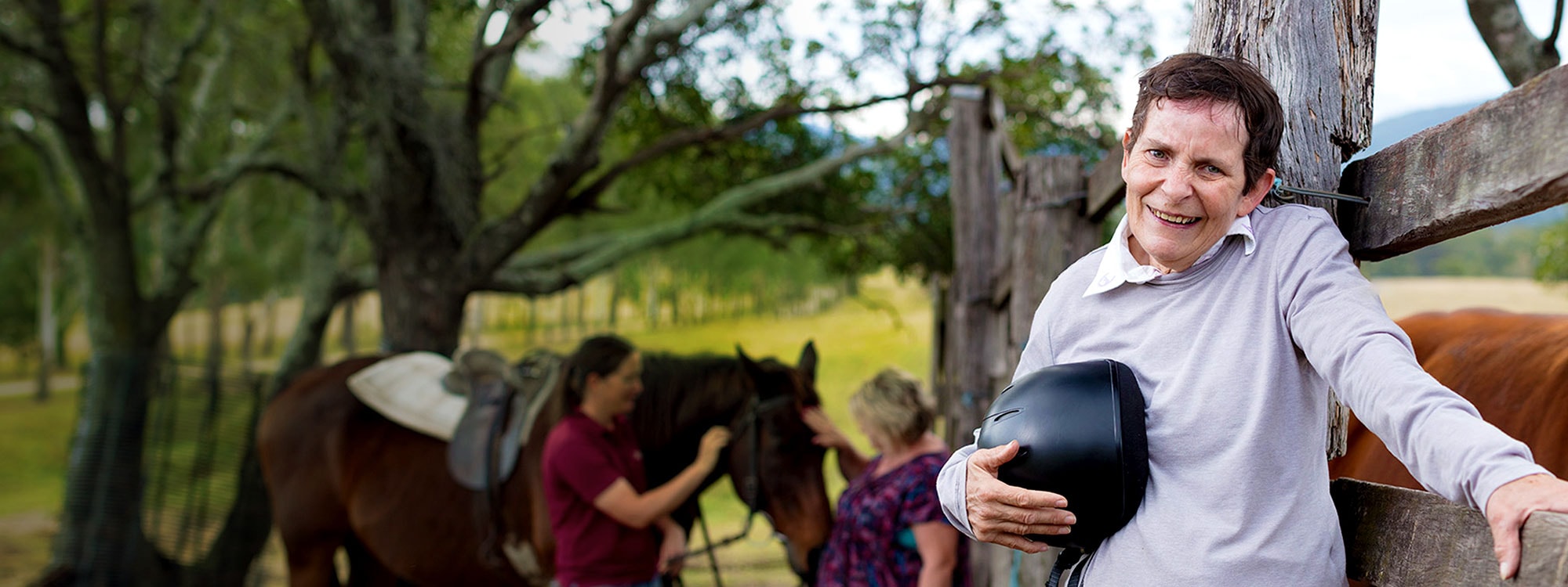 disabled man leaning against fence outdoors with staff attending a horse in the background