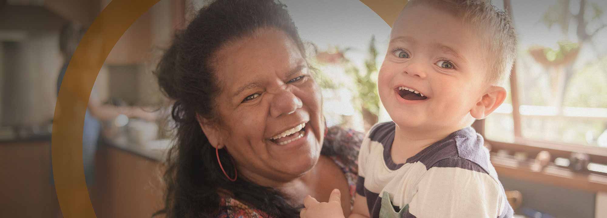 Laughing older Aboriginal woman holds up a happy little boy in her kitchen, with a young girl in the background.