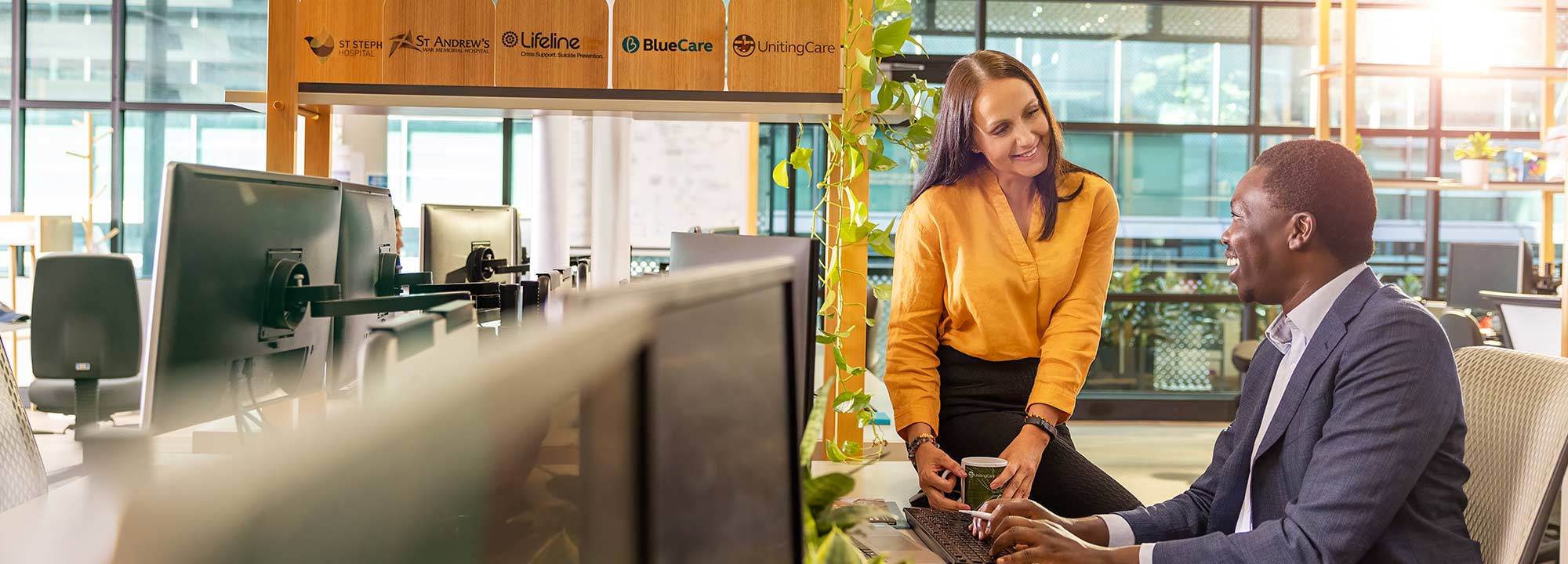 A young woman sits on a desk beside a man working at his computer in UnitingCare's head office.