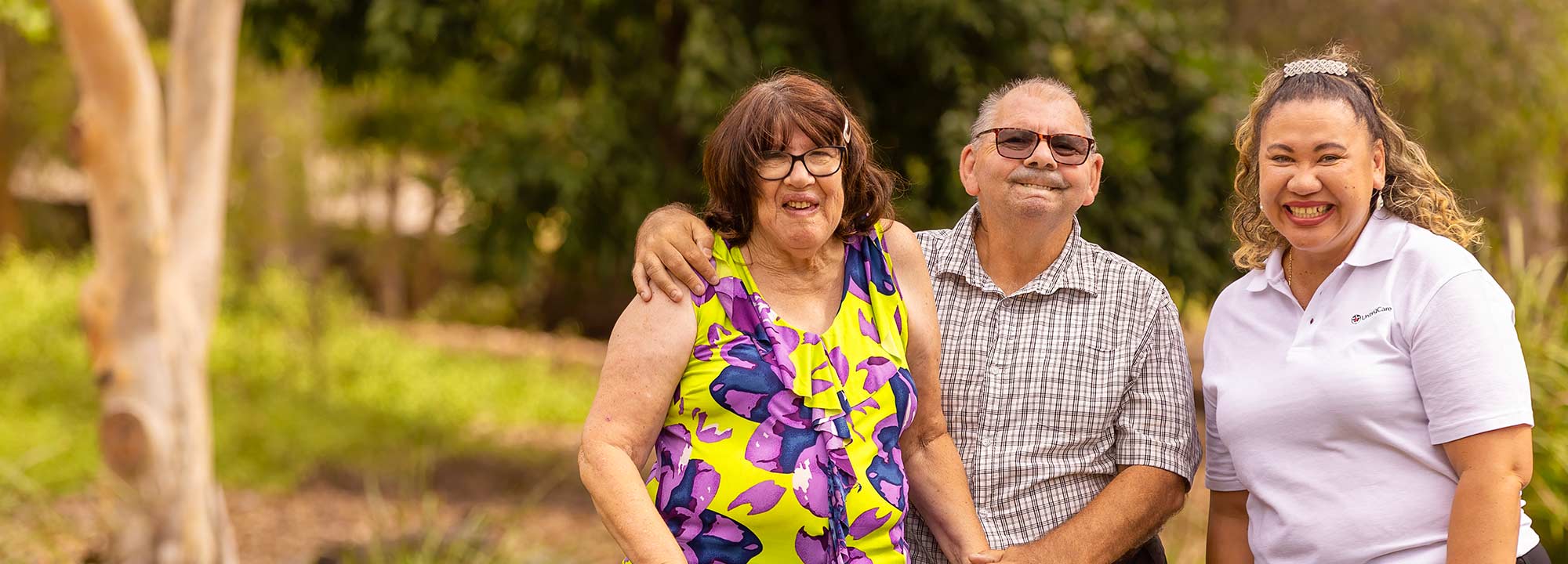 A woman using a walker stands near a park bench with a man and a UnitingCare staff member.