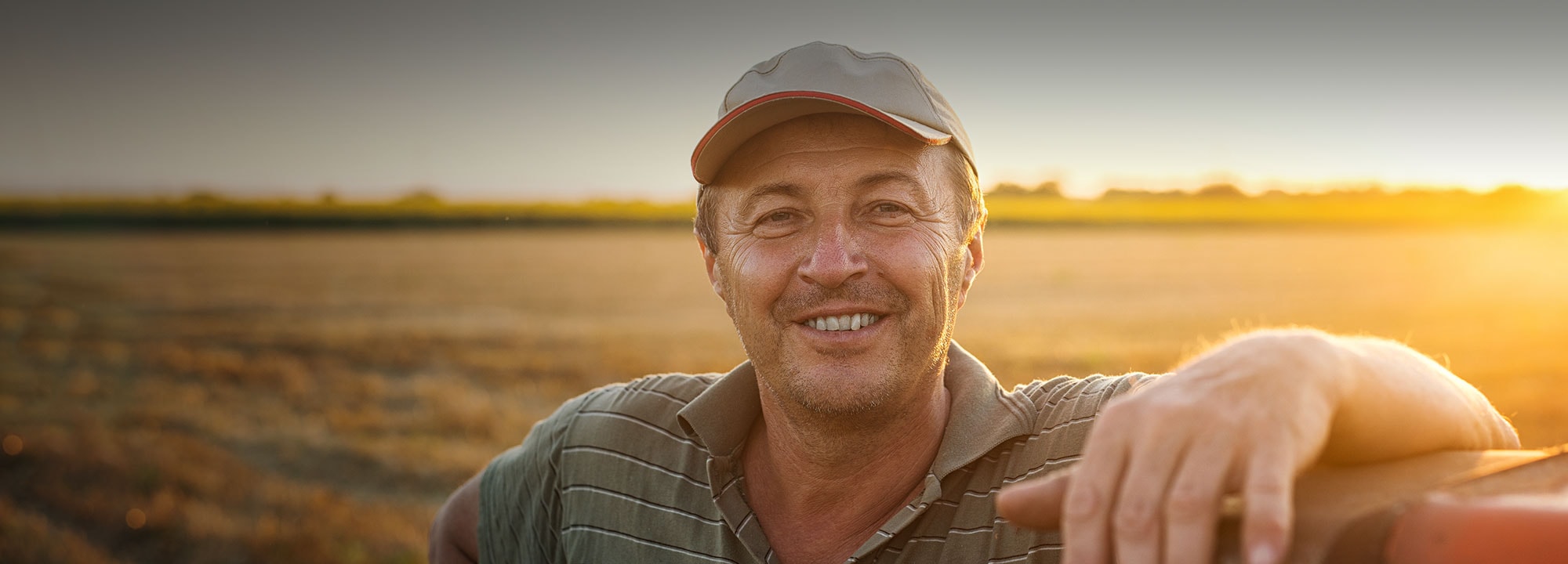 middle-aged man wearing a hat smiling towards camera in an open field as the sun sets behind him