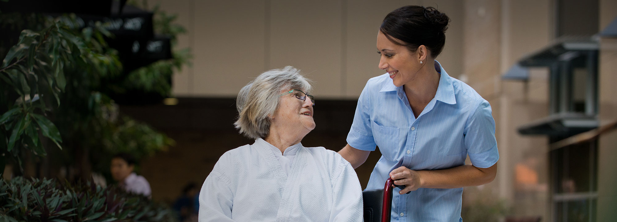 elderly woman in a wheelchair looks up and smiles at the female staff member standing beside her