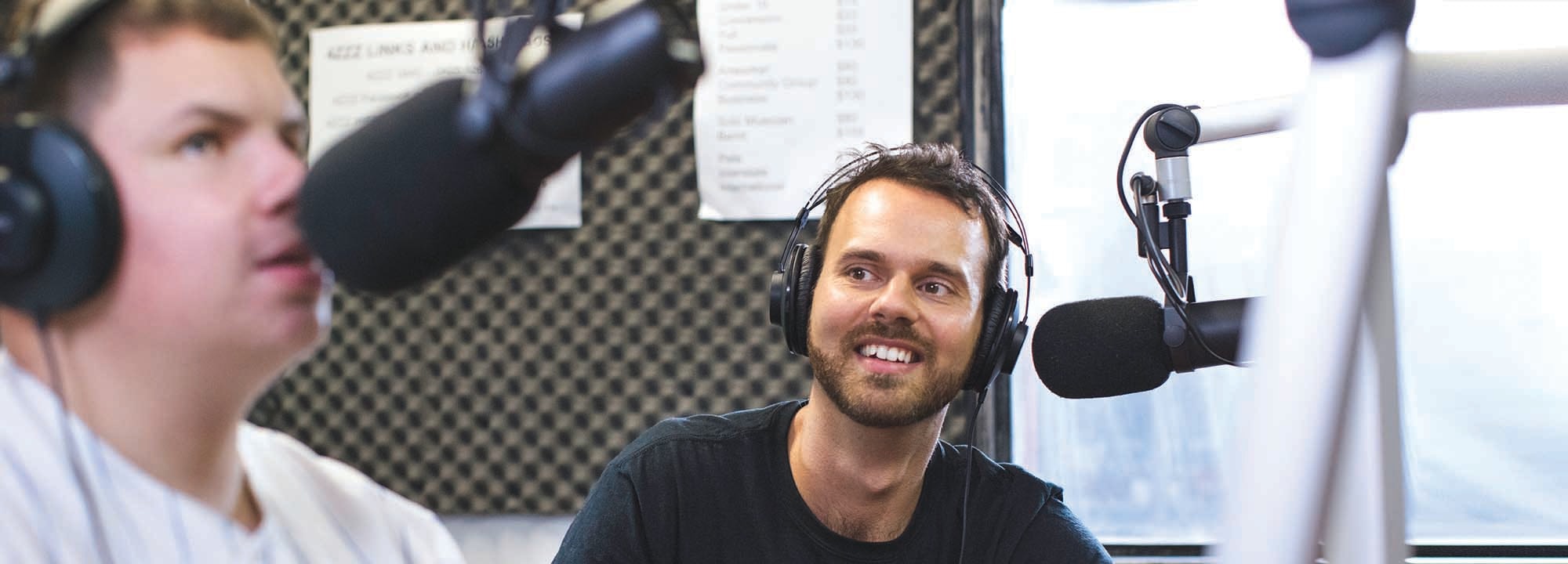 two young men sitting inside a radio studio with microphones in front of them