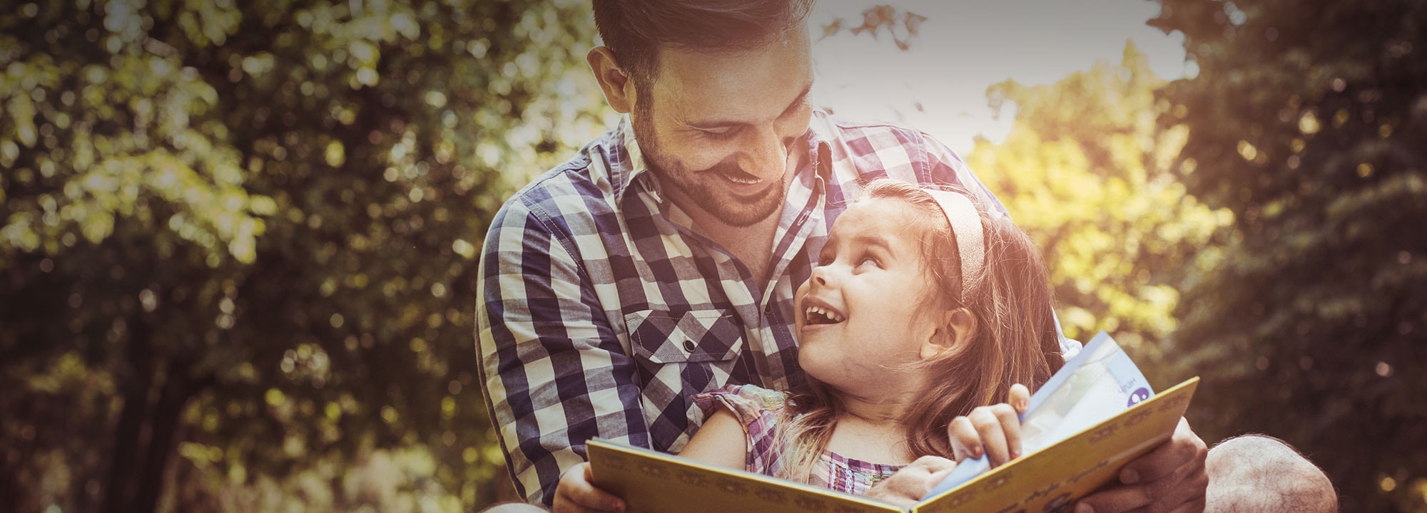a young girl sitting in her fathers lap outdoors smiling towards him as they turn the page of a book