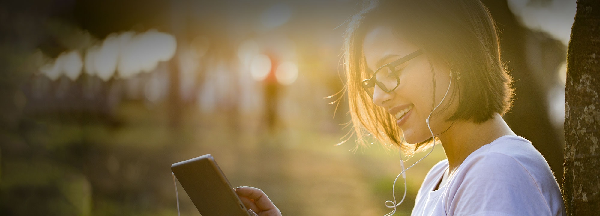 a lady sitting against a tree outdoors using an ipad