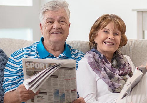 Two seniors smiling on a couch, one holding a news paper