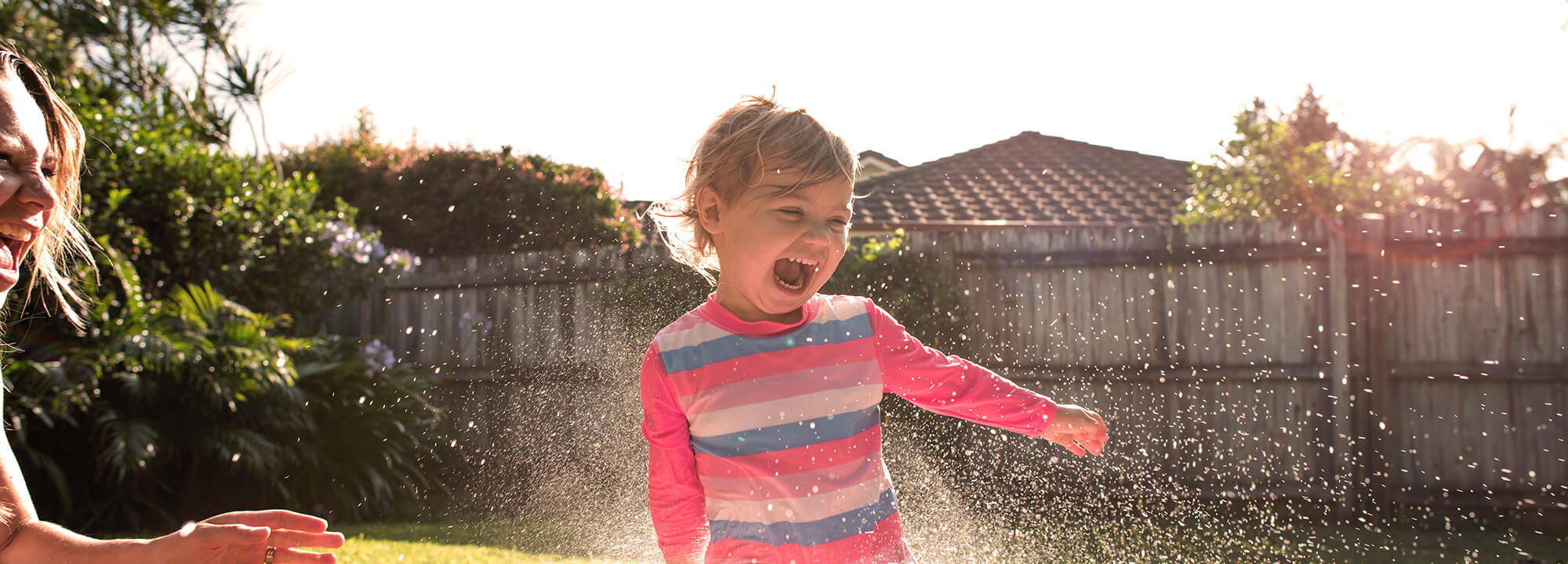 Laughing toddler splashes in a backyard blow-up pool while a woman sprays her with water from the hose.