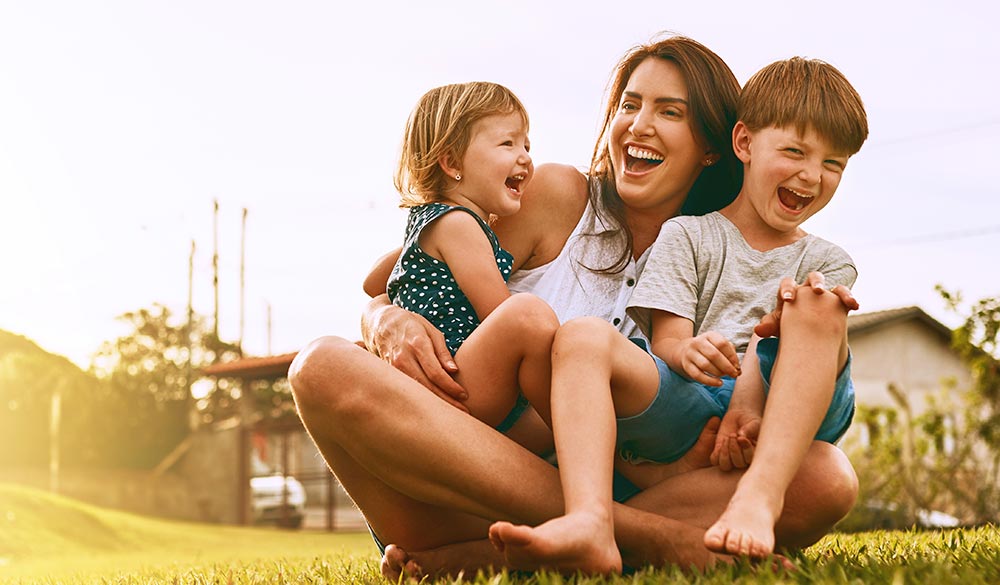 A woman with a young girl and boy in her lap sitting in parklands, laughing and smiling