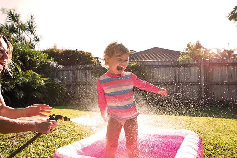 UnitingCare Early Childhood Approach A young girl in a blow up kids pool being sprayed with a hose be her mum in her back yard