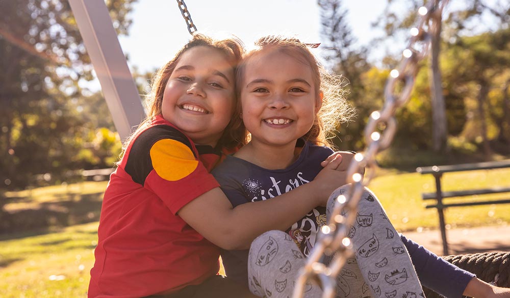 Two young girls on playground swing hugging and smiling at camera