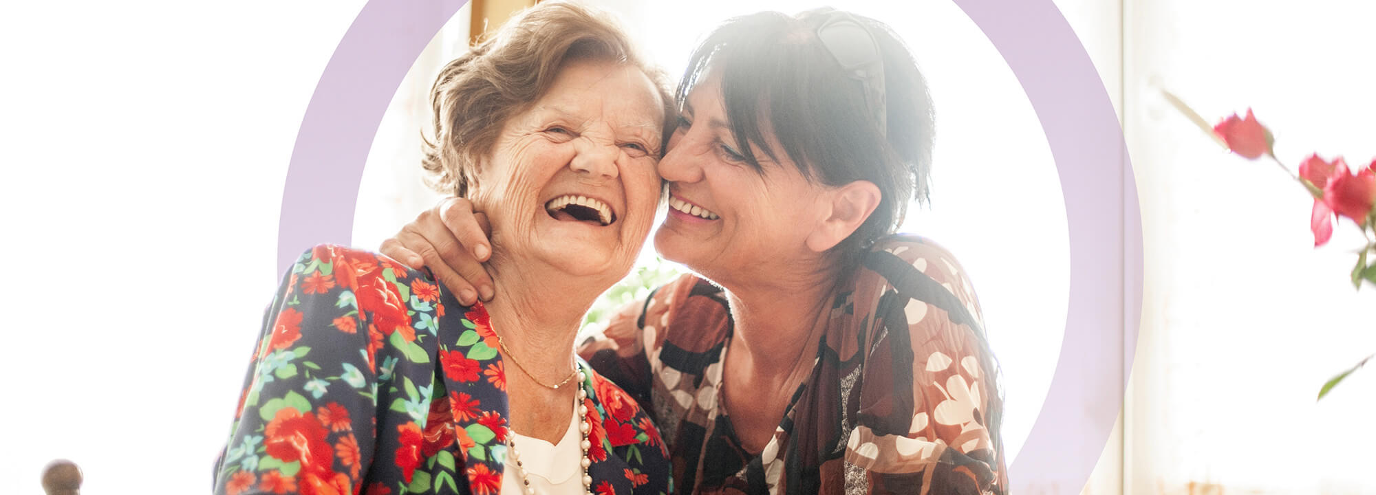 Laughing older lady sits at a table with another woman, sharing a hug and a cup of tea.