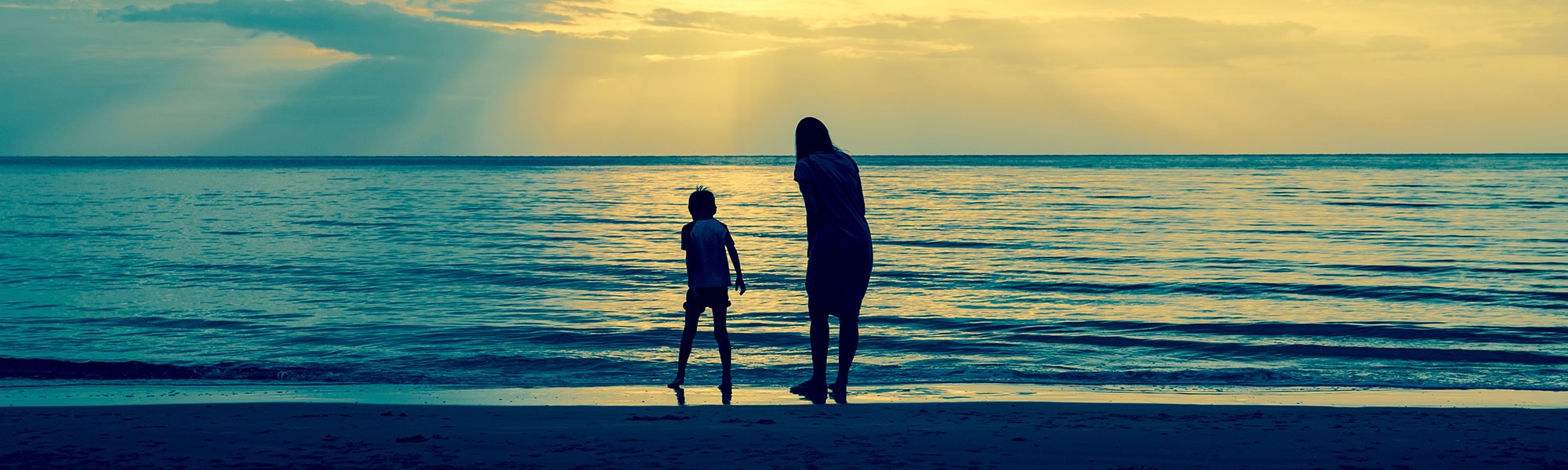 A child and adult are silhouetted against the sea as rays of light break through the clouds overhead.