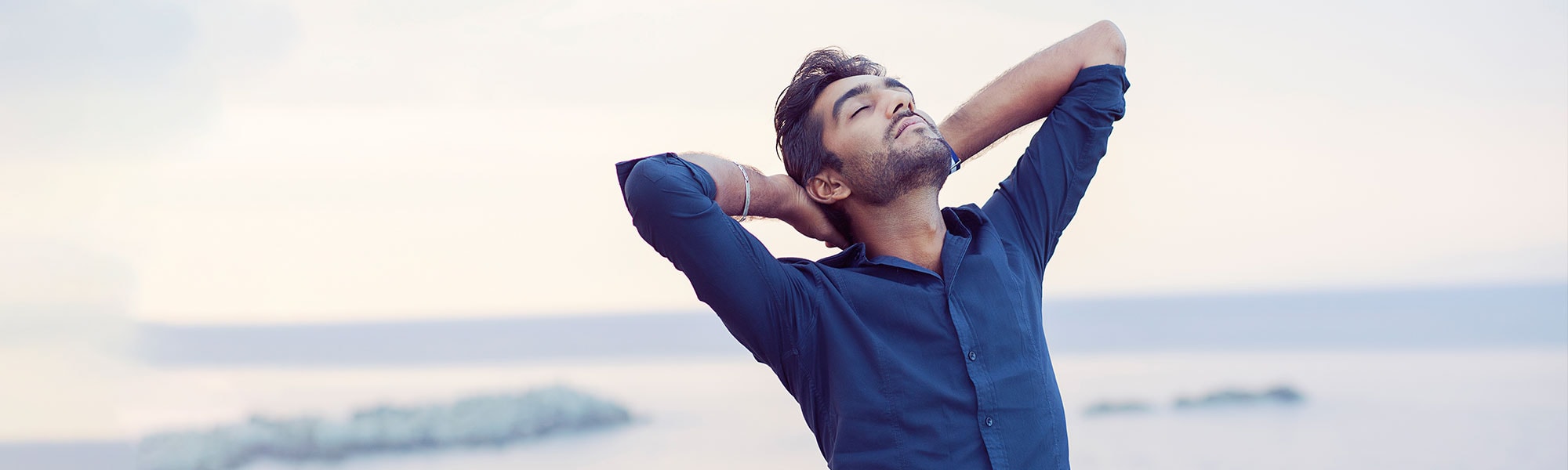 A man stretches with his arms behind his head, beside the sea.