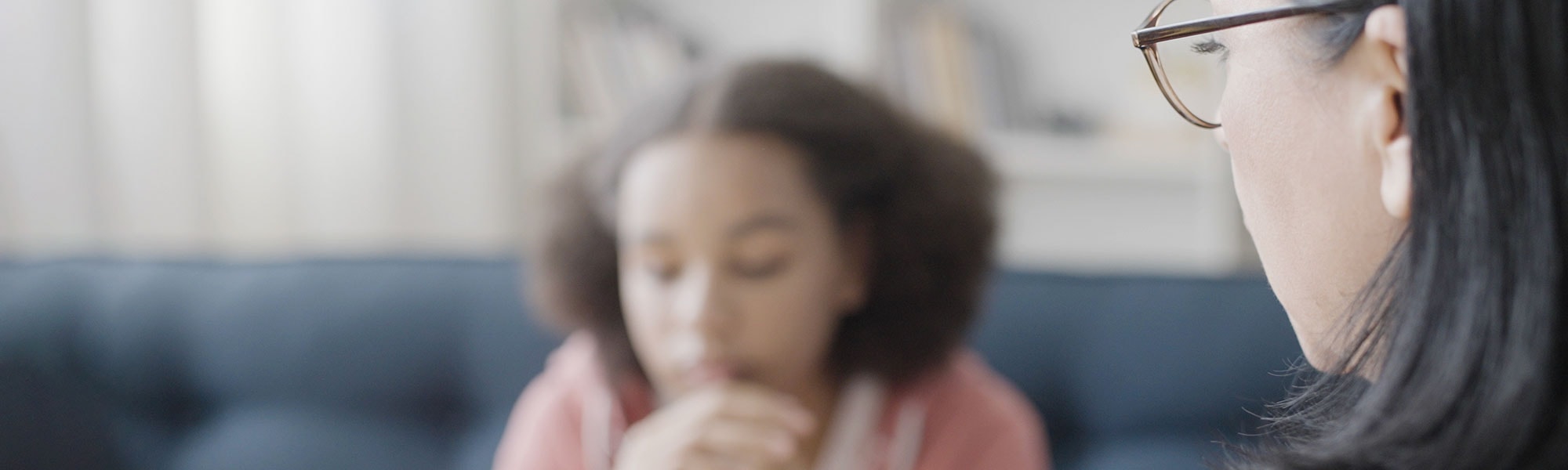 A young girl sits on a couch with her hands folded below her chin, as an adult faces her.