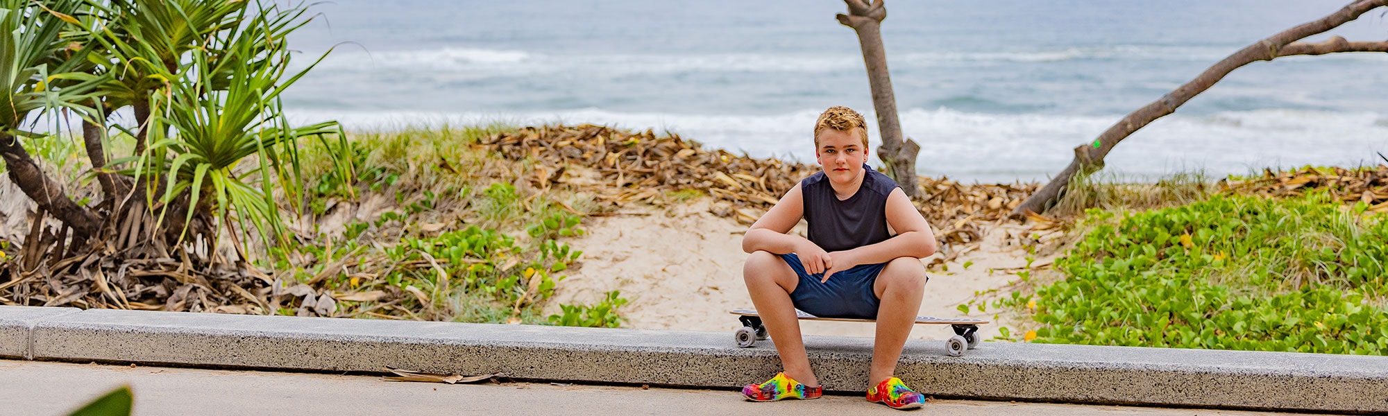 A young man sits in the gutter with his skateboard, with a tree and the beach behind him.