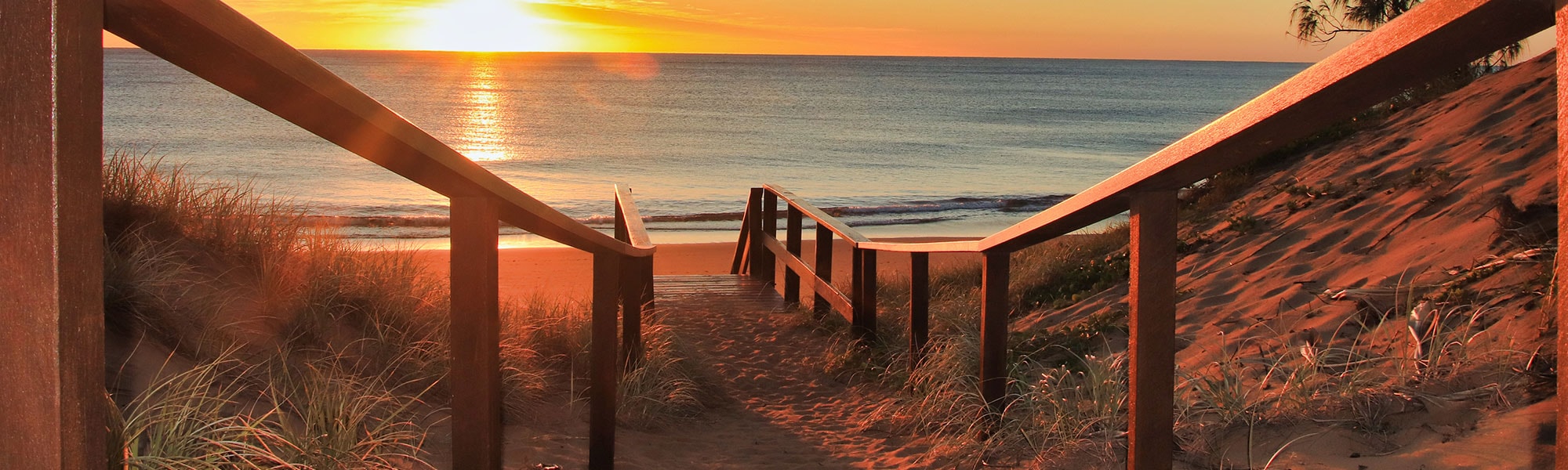 A sandy beach path stretches towards the ocean as the sun sets.