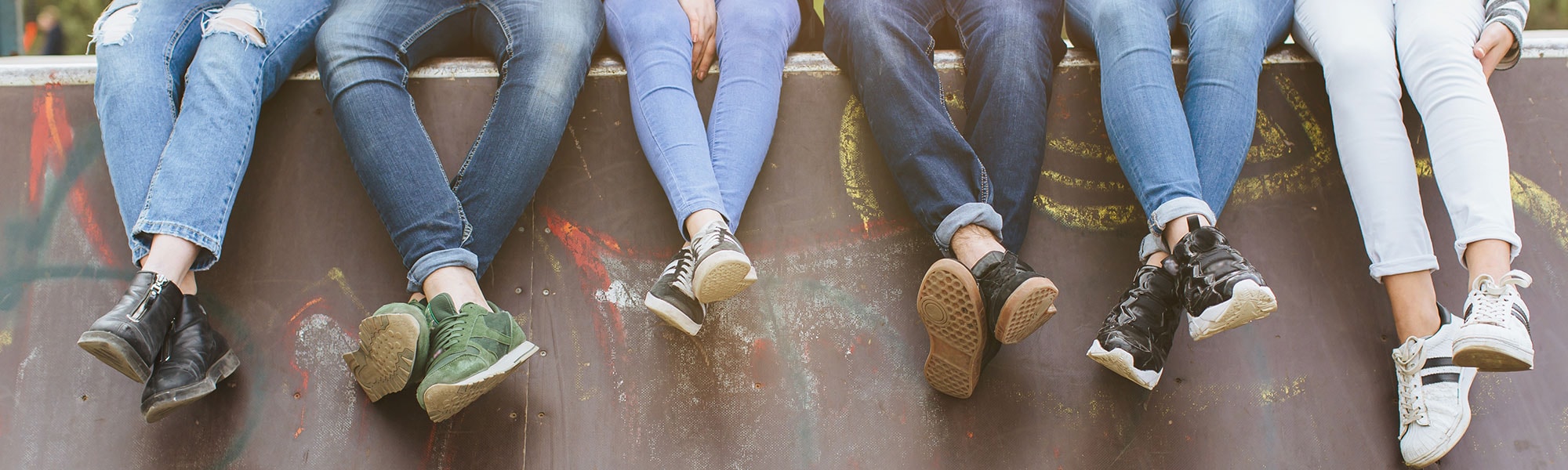 A group of unidentified teens wearing jeans and sneakers sit on a wall.