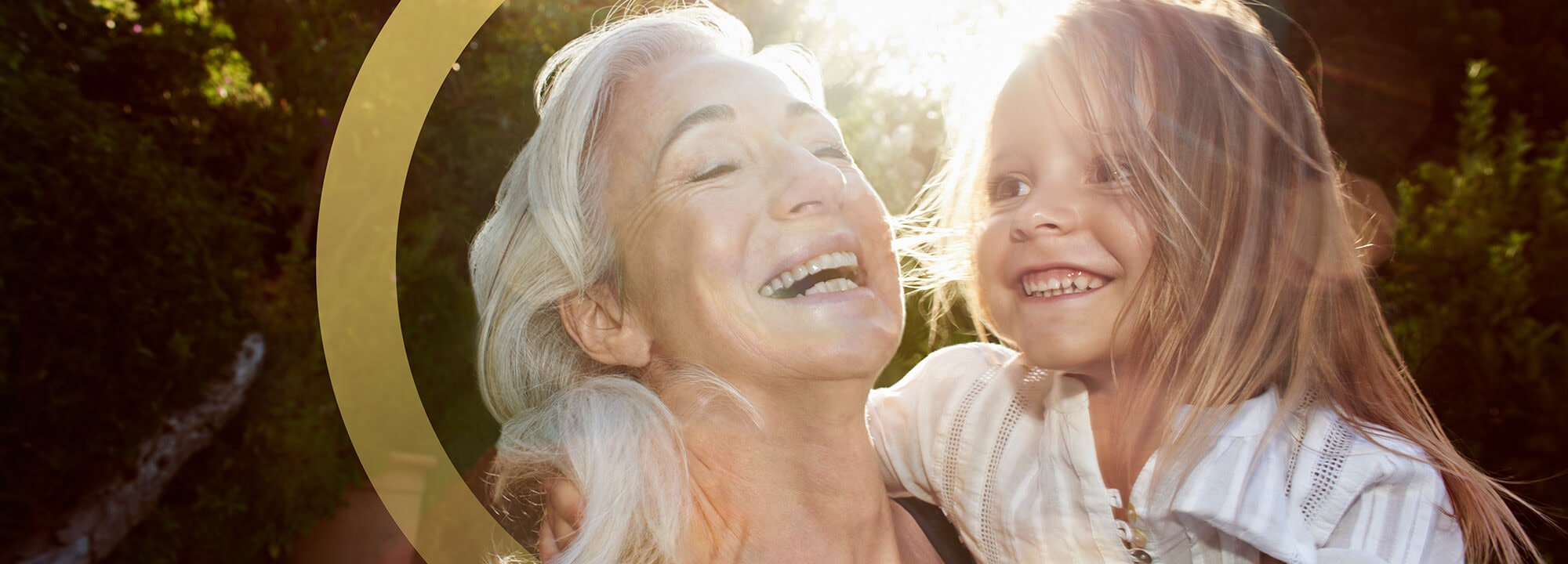 Laughing older woman with silver hair holds a smiling young girl in the garden.