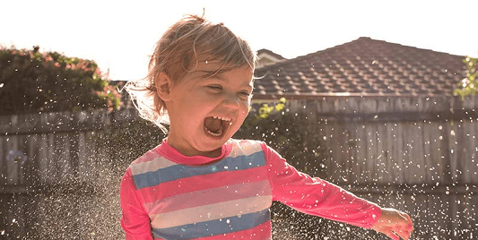 NDIS Early childhood approach CTA A young child in a stripey top laughs with joy while playing in a backyard sprinkler.