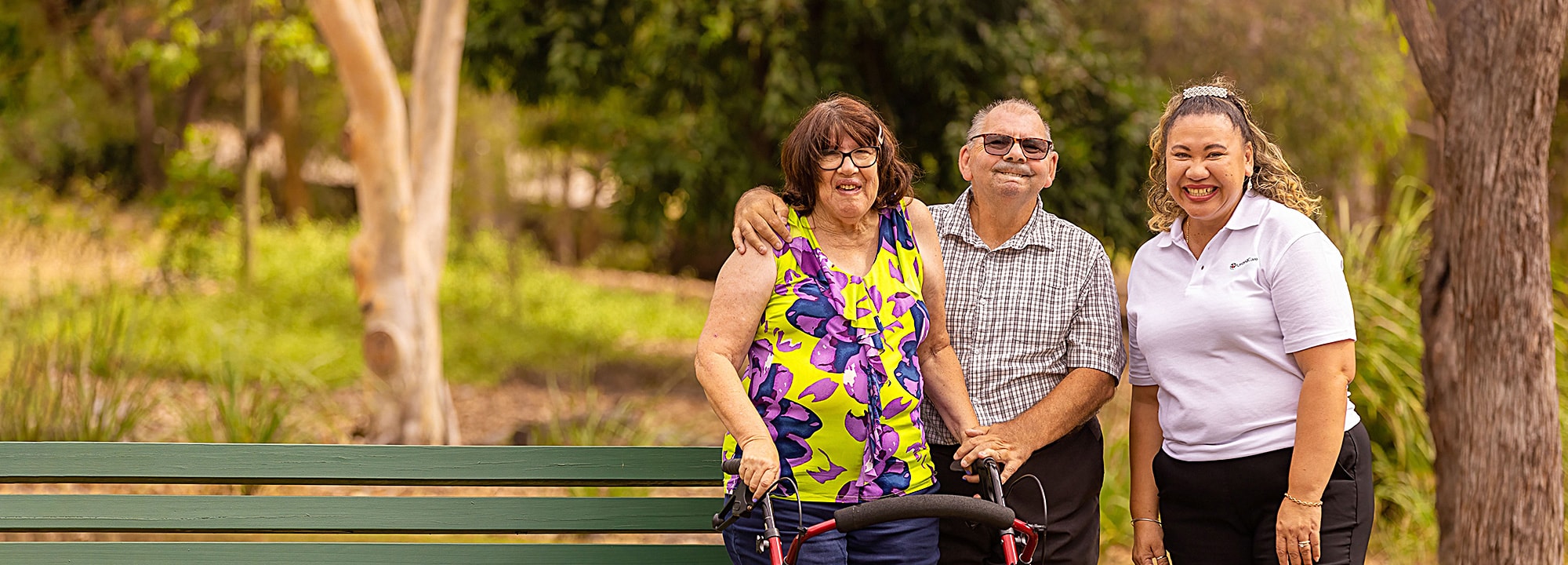 A woman who uses a wheelie walker stands with her partner and a UnitingCare disability support worker beside a bench in a country park.