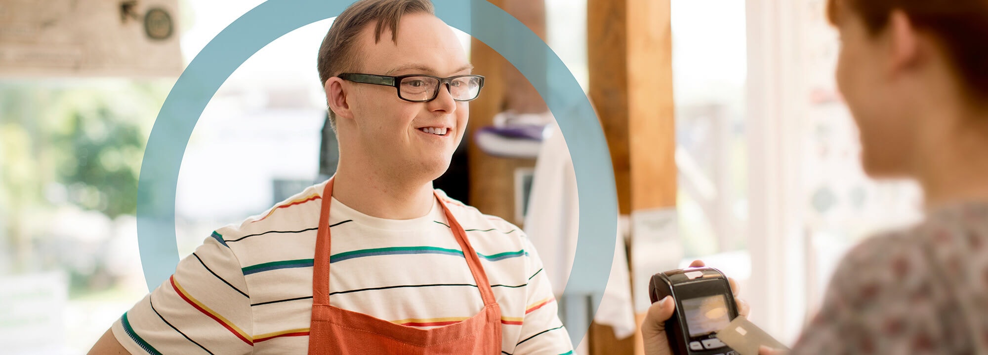 Young man with disability, wearing an apron, holds up an EFTPOS machine to a customer at the store where he works.