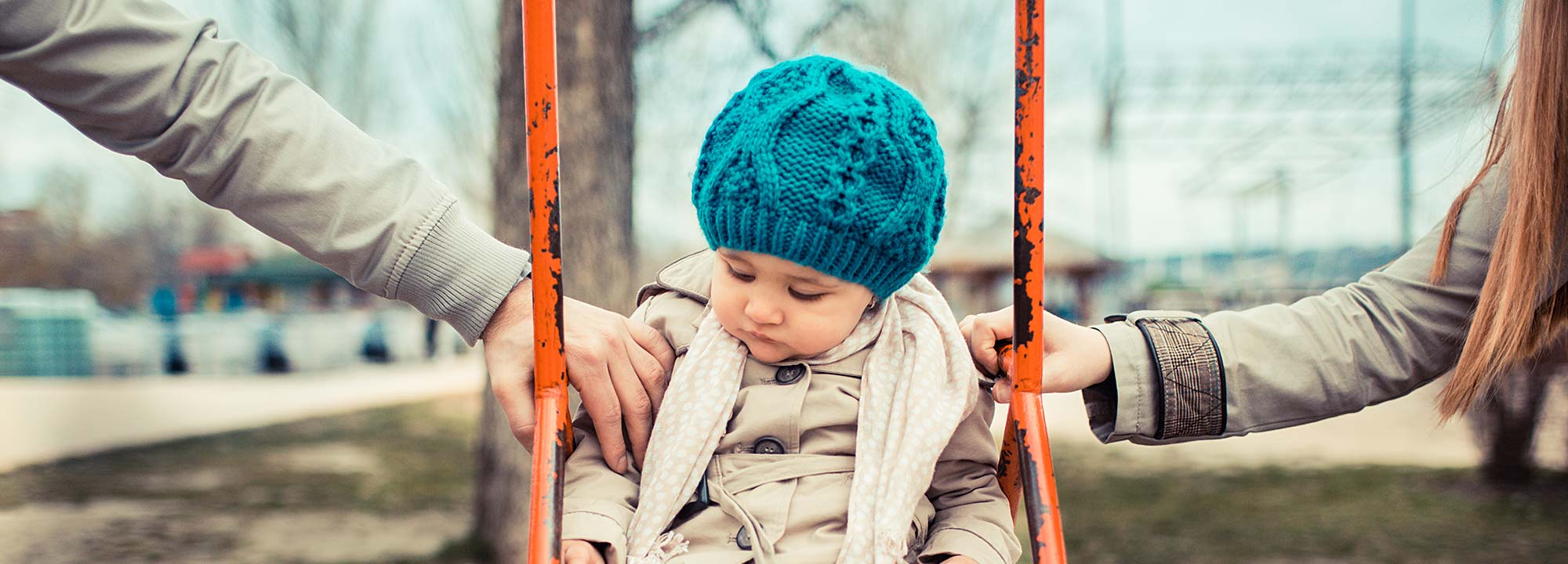 A young child in a beanie plays on a swing with a parent on either side.