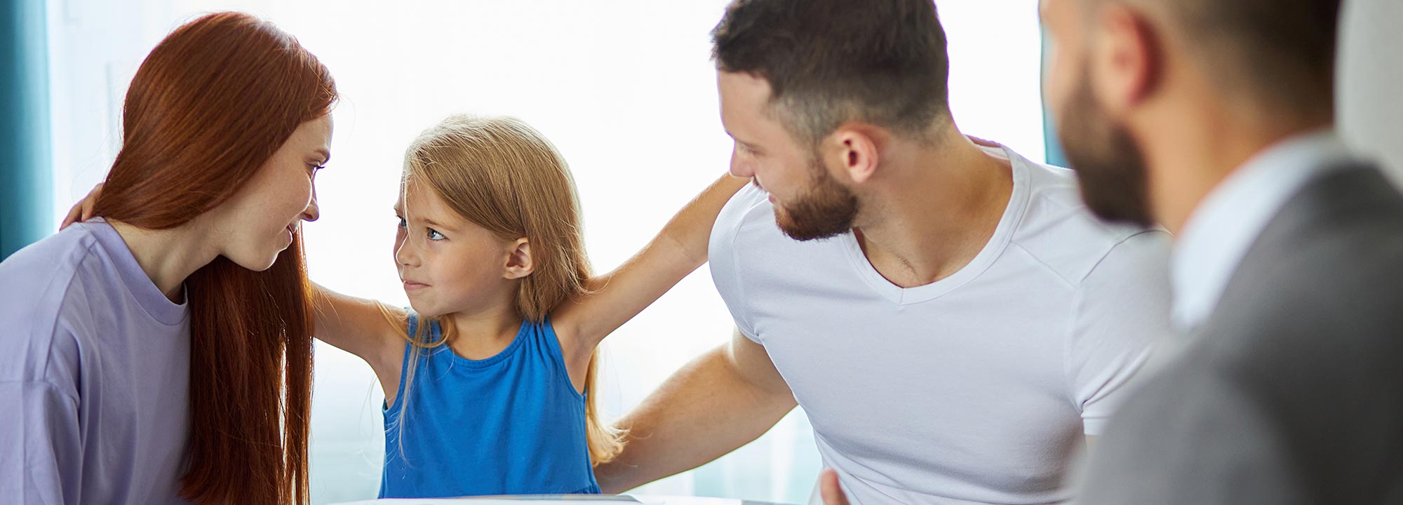 A young girl stands between her mother and father, who are sitting at a table with a mediator.