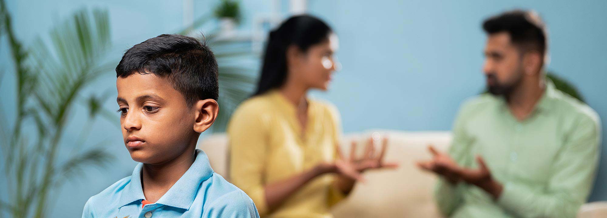 A young boy holding a teddy bear looks sad as his parents argue in the background.