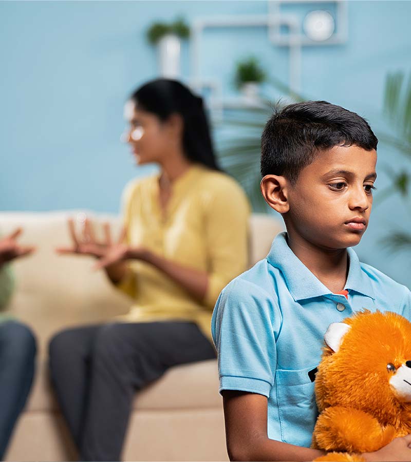 A young boy holding a teddy bear looks sad as his parents argue in the background.