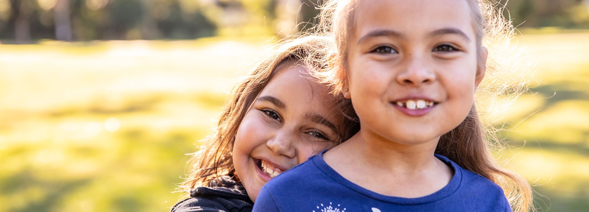 A smiling young girl hugs another smiling young girl in a park.