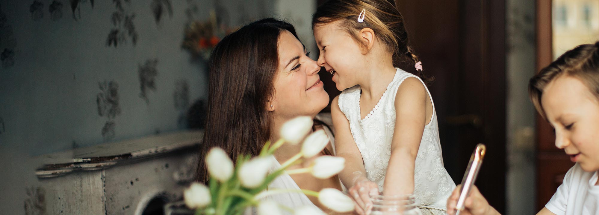 A mother and child bumping noses and smiling at each other beside another child drawing